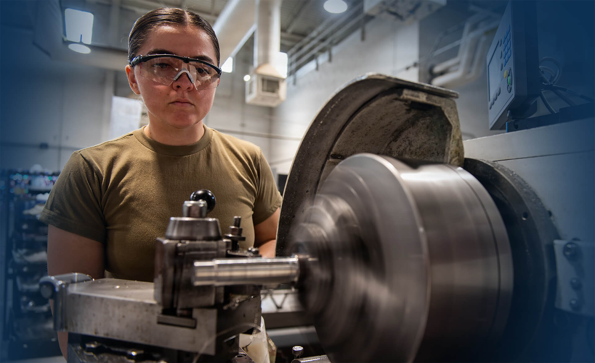 A technician wearing safety glasses operates a metal lathe in an industrial workshop, carefully machining a rotating metal component while monitoring the equipment controls.