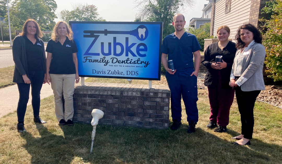 Five people stand outside next to a blue sign for Zubke Family Dentistry, which reads “Zubke Family Dentistry — The Key to a Healthy Smile, Davis Zubke, DDS.” The group is smiling, and two people are holding tumblers. The sign is mounted on a short brick base in front of the dental office.