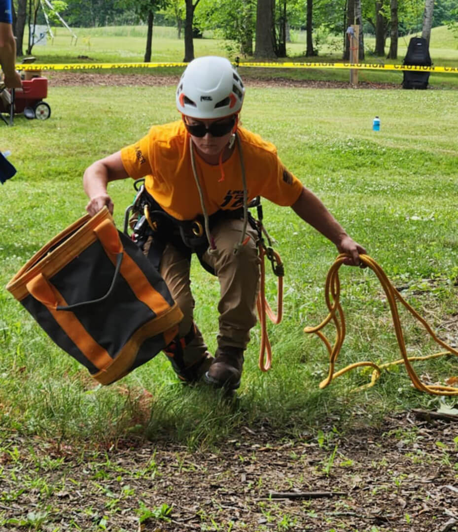 Hanna, a Line Clearance Tree Trimmer, wearing safety gear with a helmet and harness, crouching on the grass while carrying a gear bag in one hand and holding a climbing rope in the other.