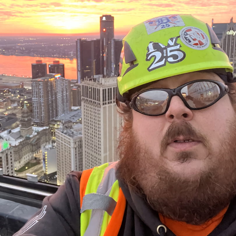 Charles — an Ironworker Registered Apprentice — wearing safety gear, including a bright yellow hard hat and reflective vest, while taking a selfie high above Detroit at sunrise. The GM Renaissance Center and other downtown skyscrapers are visible in the background, with the Detroit River glowing orange from the rising sun.