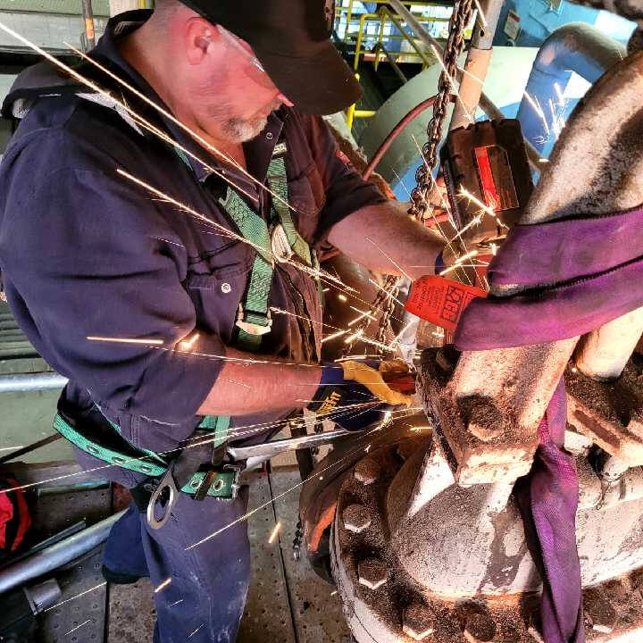 Todd, a Registered Stationary Engineer Apprentice at Nexteer Automotive, wears safety gear — including safety glasses, a harness, gloves, and a cap — while using a power tool on a large flanged metal component, producing sparks in a heavy industrial environment.