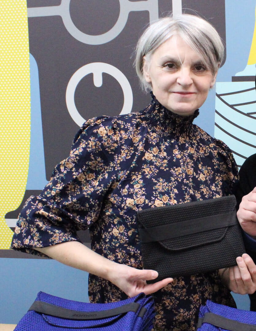 Mariya, a Ukrainian refugee and sewing machine operator at Zaman International who participated in the Michigan Works! PATH program, wearing a navy blue floral blouse and holding a small black textured clutch-style pouch.