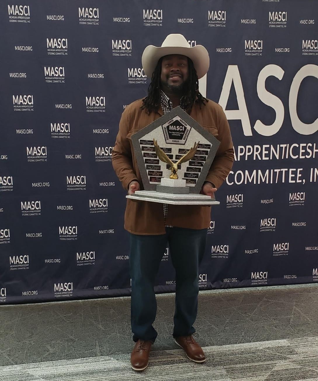 Anwar, a Black man in a cowboy hat, holding a large award and standing in front of a MASCI banner.