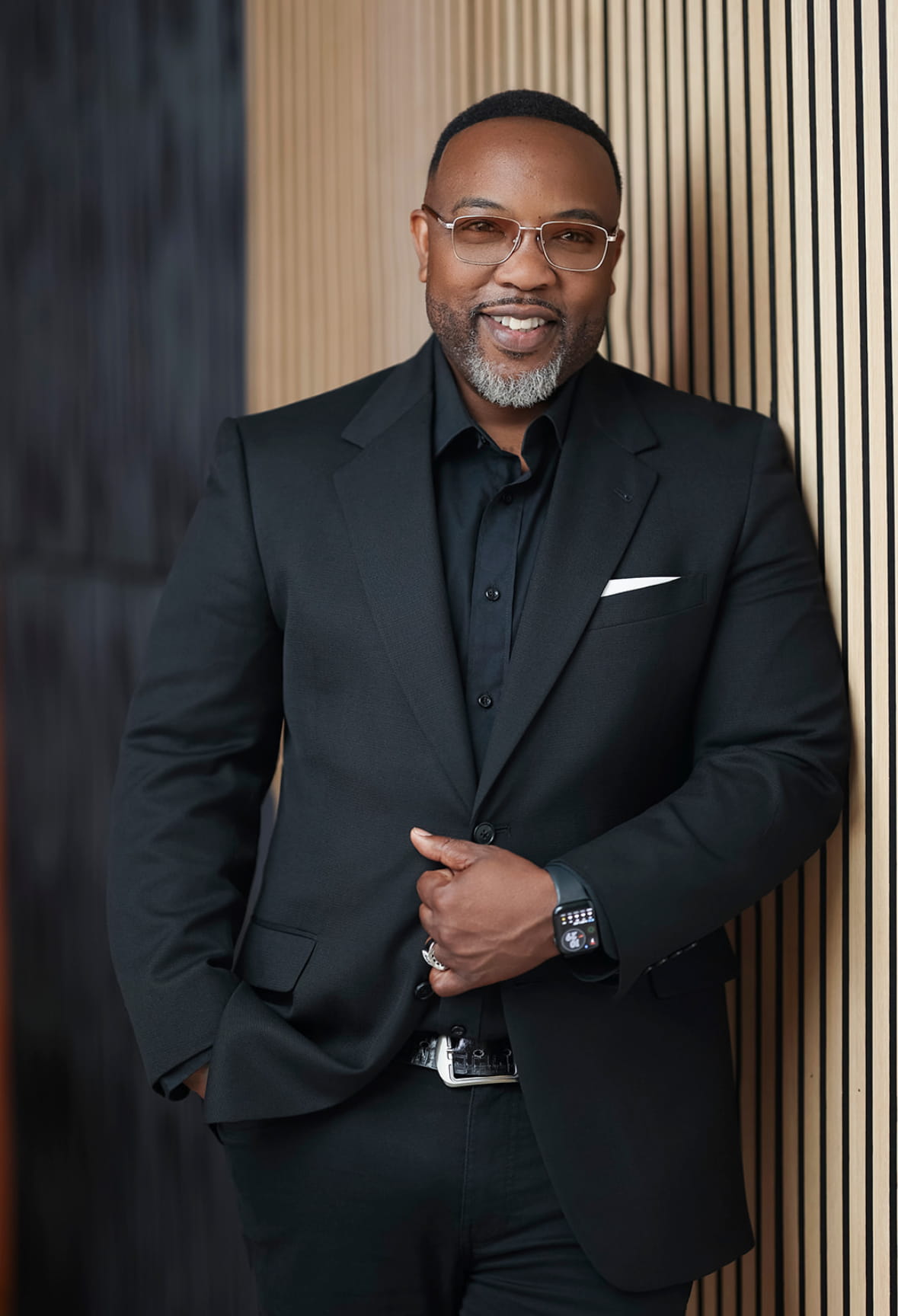 Gerod, a Black man wearing a black suit and glasses and leaning against a wooden wall