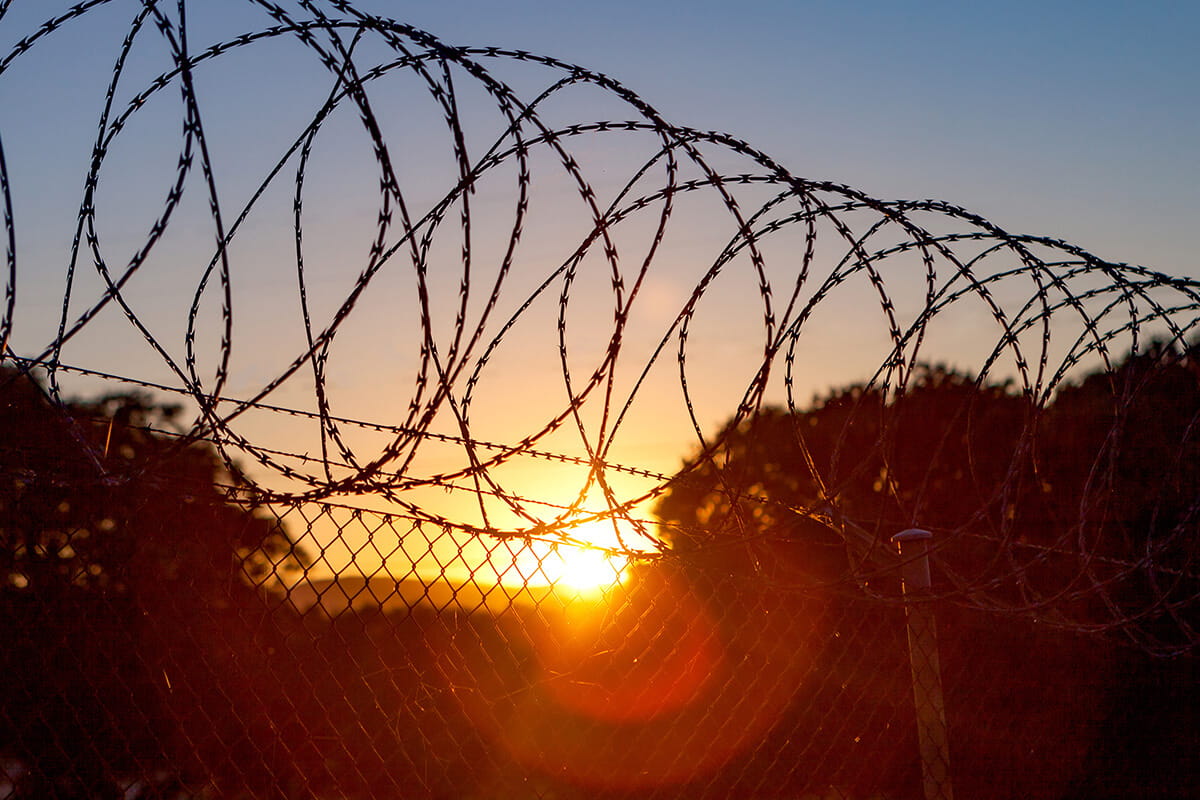 Silhouette of chain-link fencing topped with barbed wire at twilight.