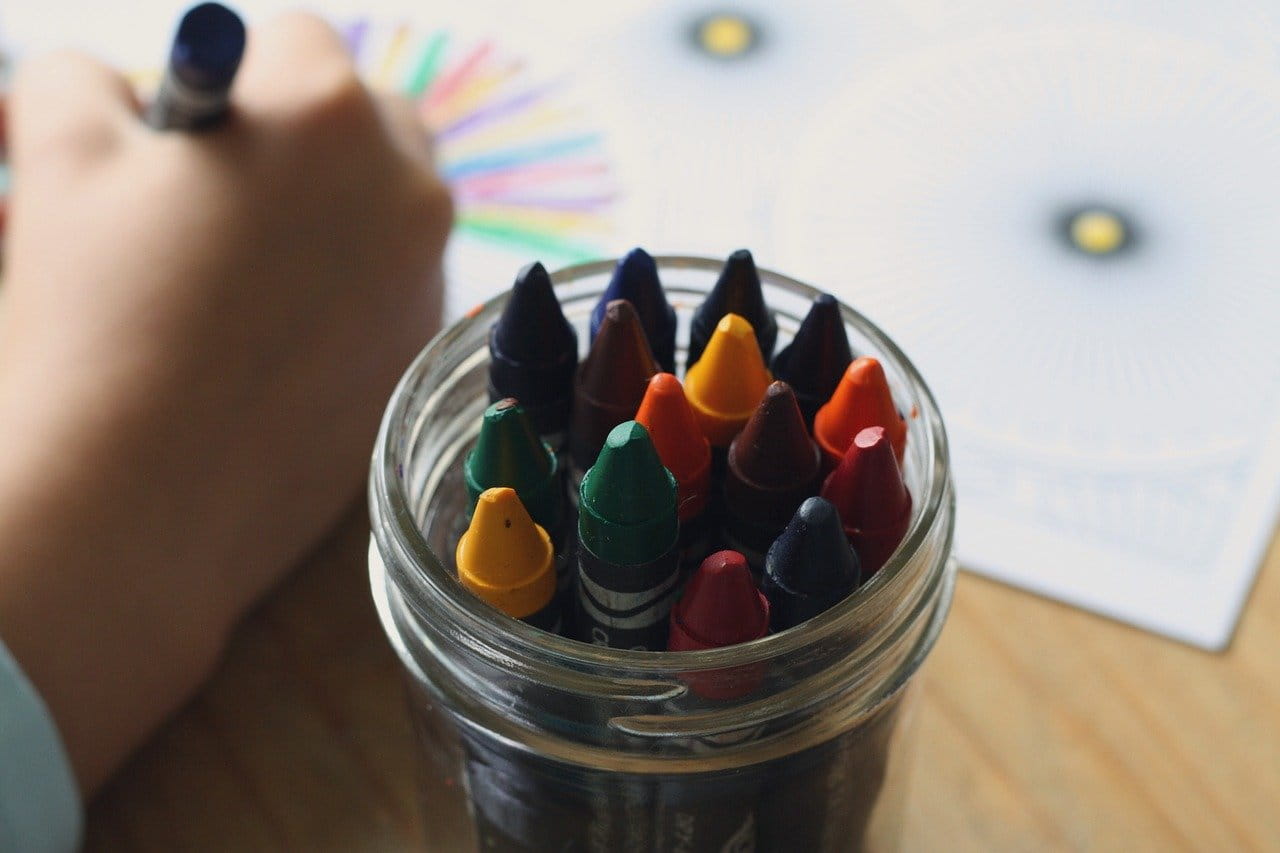 Close up of crayons in a glass container with a child's hand coloring next to them.