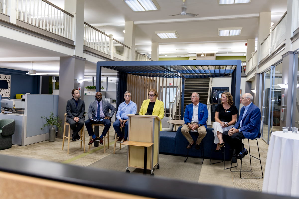 A group of 6 sit on stage with Director Corbin presenting at a podium front and center of them