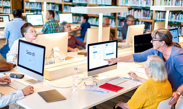 A diverse group of people in a library using computers, with a woman assisting another at a desk. Bookshelves are visible in the background.