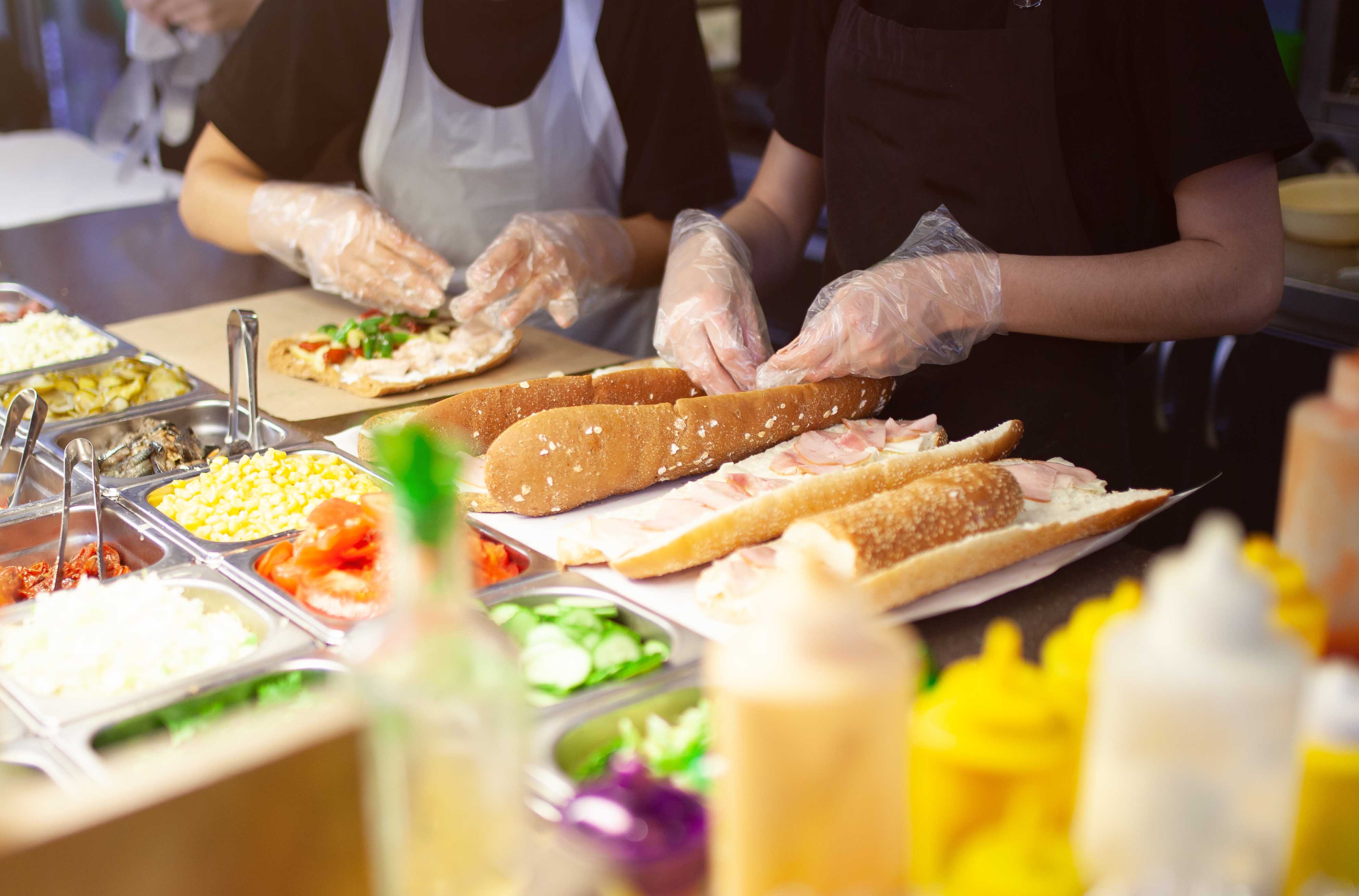 gloved hands putting toppings on sub sandwiches 