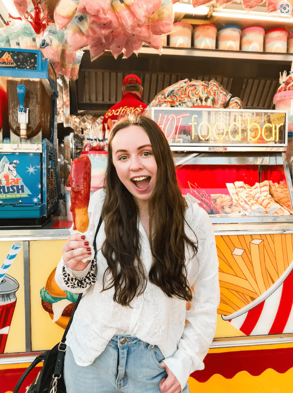 Woman in front of a festival stall eating a hotdog on a stick and smiling