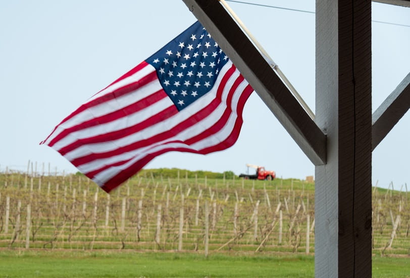 A US Flag in front of a farmhouse. You can see the field is near harvest time.