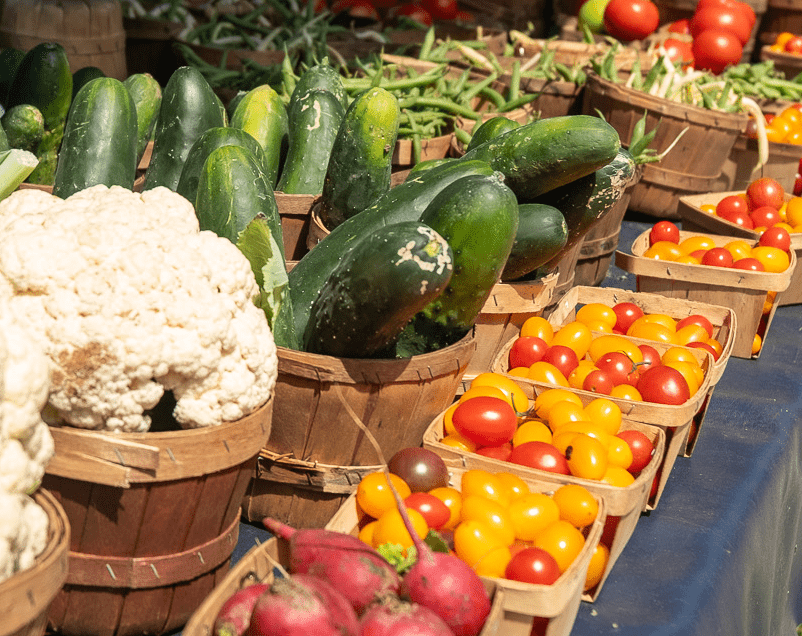 Brightly colored vegetables ready for sale at a farmers market: cauliflower, cucumbers, tomatoes and peas.
