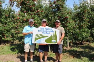 Three men in an orchard holding a sign that says 