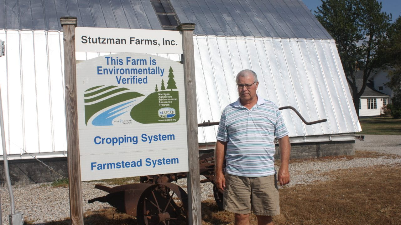 Stutzman standing in front of farm equipment with a sign that says "This Farm is Environmentally Verified: