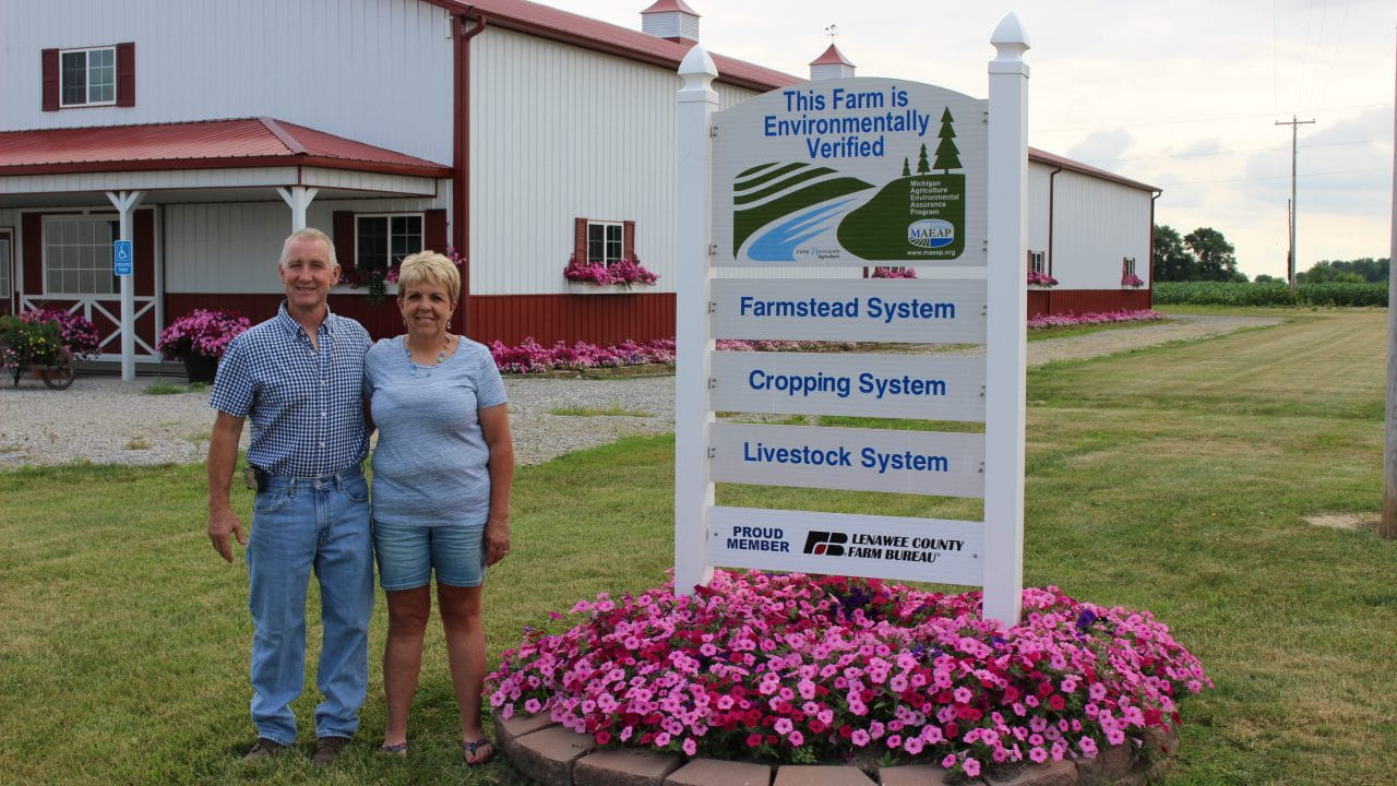 Matt and Kelly Carpenter standing in front of a mound of flowers with a MAEAP sign that indicates they have verified their farmstead, cropping system and livestock system