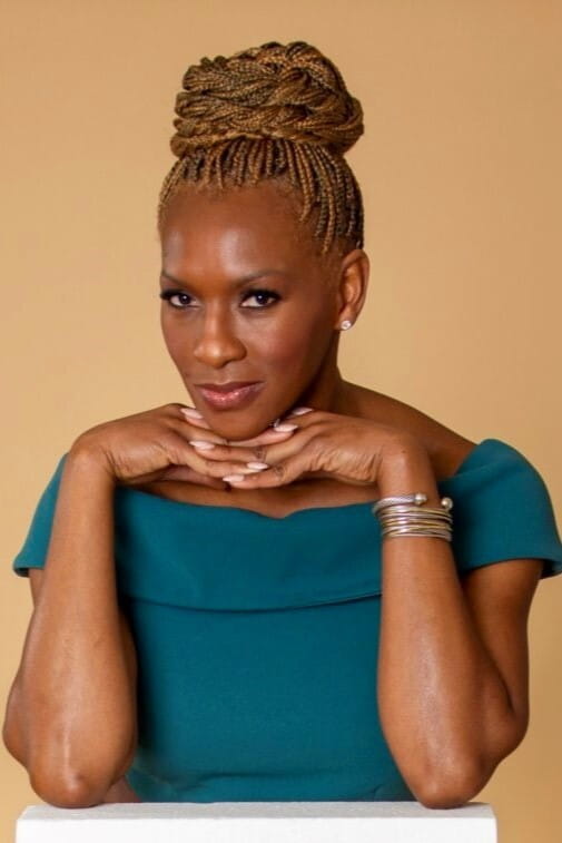 Claudia looks at the camera, smiling with her hands resting on her hands. She wears a teal top and is set against a beige background. Her hair is in a braided blond bun.