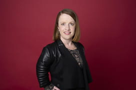 Rebecca looks at the camera smiling. She wears a black blazer over a black top with her hands on her hips. She is set against a dark red background.