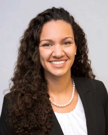The image is a headshot portrait of a young woman with a warm, inviting smile. Her hair is dark brown, thick, and styled in loose, bouncy curls that fall past her shoulders. Her complexion is fair with a warm undertone with hazel eyes. She is wearing professional attire. A classic black blazer is visible, open to reveal a crisp white top with a pleated neckline pearl necklace.