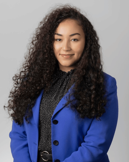Annika Clemens is a biracial Black woman with a light skin tone and long, curly dark brown hair. She is wearing a blue blazer and a black and white polka-dot blouse as she smiles at the camera with her arms crossed. She stands in front of a plain grey background.