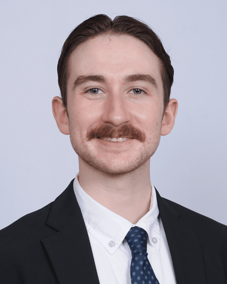 David O’Keefe is a white male with a light skin tone, short brown hair, and a mustache. He is wearing a black suit, white shirt, and blue tie. He is photographed in front of a plain grey background.