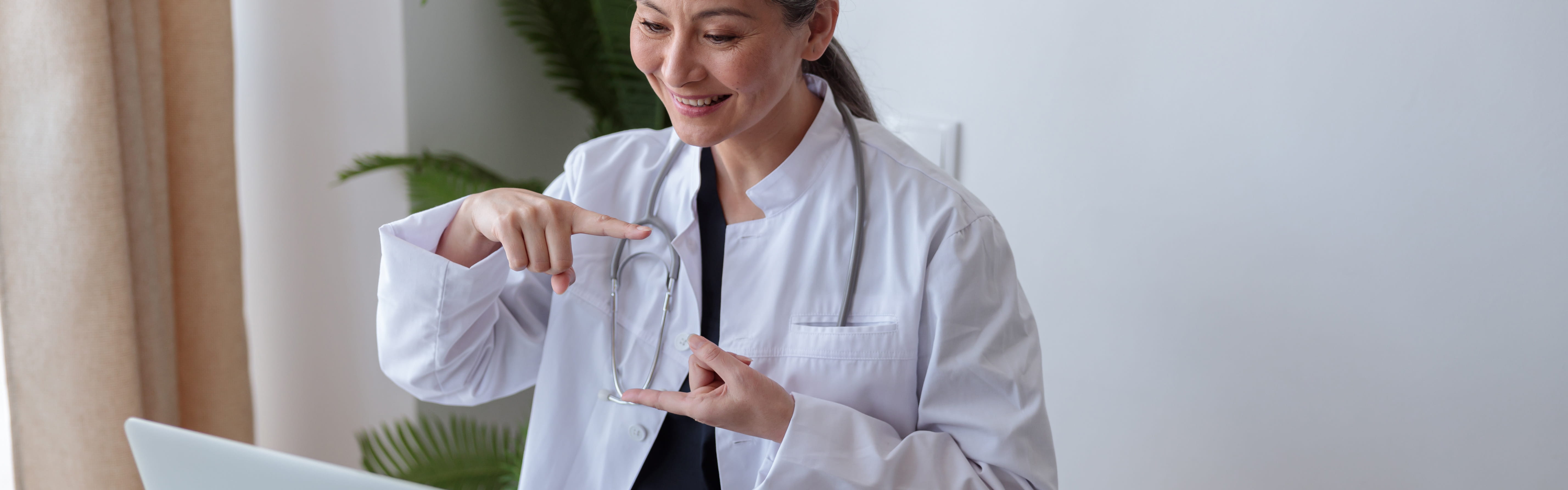 A healthcare professional wearing a white lab coat and a stethoscope signing in American Sign Language while seated at a desk with a laptop.