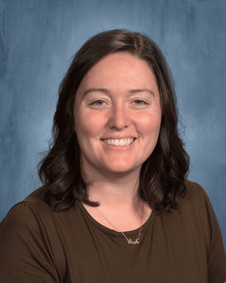 Emma Heisey is a white woman with light skin and shoulder-length curly brown hair. She is wearing a brown short-sleeve shirt and is photographed in front of a plain grey-blue background.
