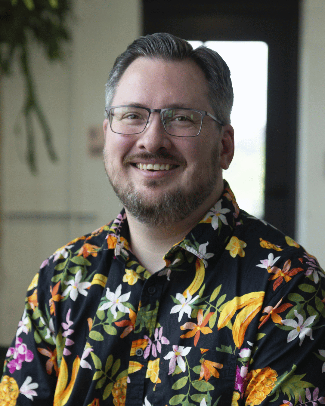 Eric Welsby is a white male with short greying dark brown hair, a beard, and rectangular glasses. He is wearing a black button-up shirt with a colorful tropical floral pattern. He is photographed in an indoor setting with natural light and greenery. 