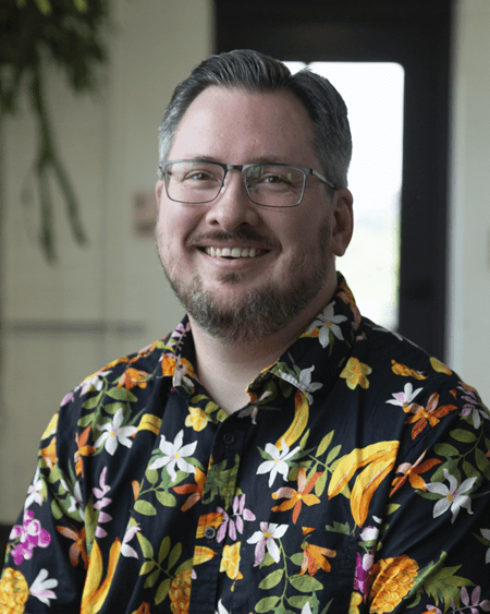 Eric Welsby is a white male with short greying dark brown hair, a beard, and rectangular glasses. He is wearing a black button-up shirt with a colorful tropical floral pattern. He is photographed in an indoor setting with natural light and greenery.
