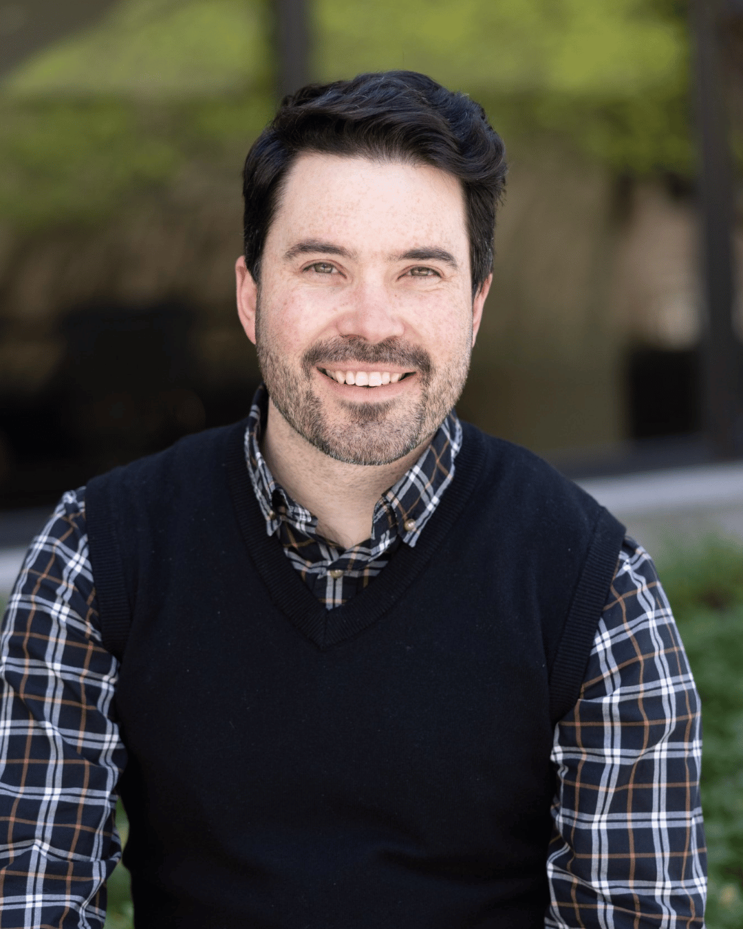 James is a white male with short black hair and a trimmed black beard. He is wearing a black vest over a plaid patterned dress shirt. He is photographed in front of an outdoor garden with leafy plants and a tree. 