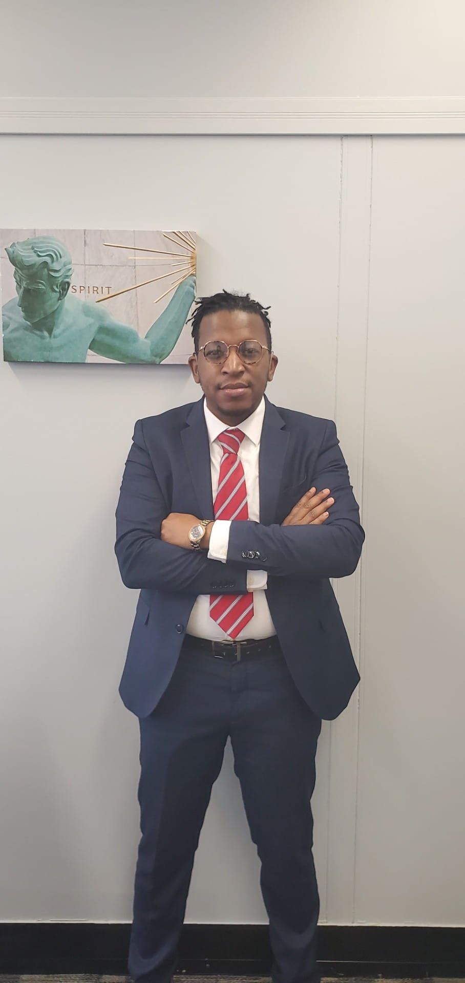 Image of a man with short black hair and dark skin in a navy blue suit, white shirt, and red striped tie standing with his arms crossed.