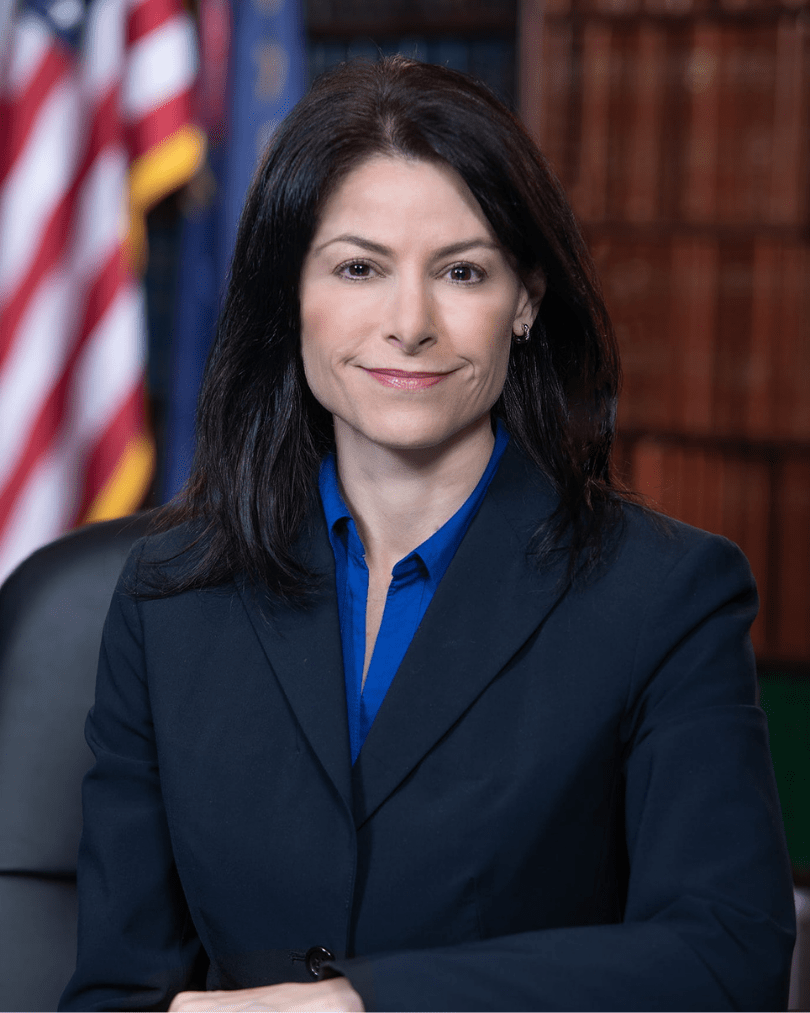 Dana Nessel, Michigan Attorney General: She has dark, shoulder‑length hair wearing a dark blazer and blue shirt, seated in front of blurred bookshelves and the Michigan and U.S. flags.