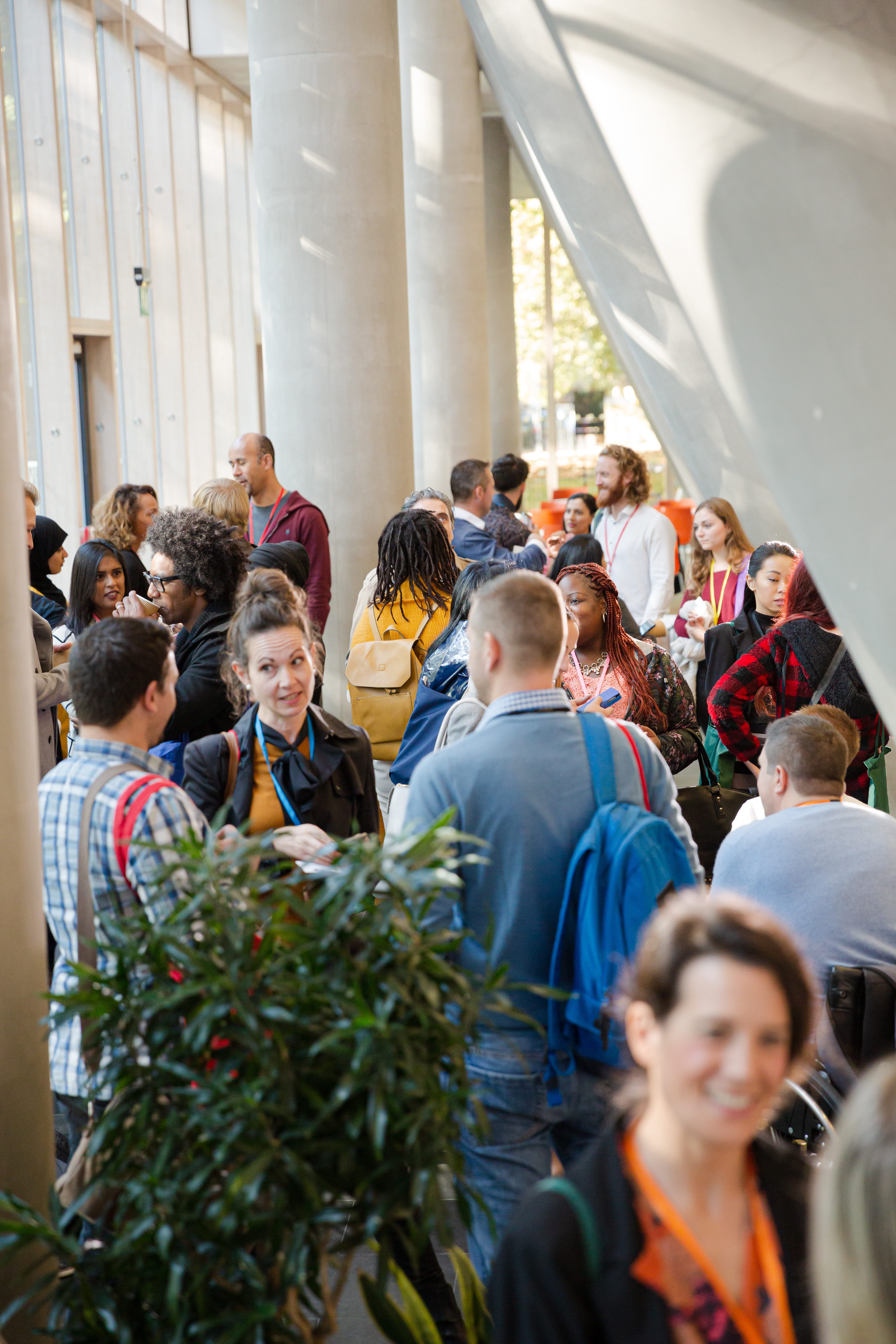 A crowd of people wearing event badges gather and talk in a bright lobby with tall columns.