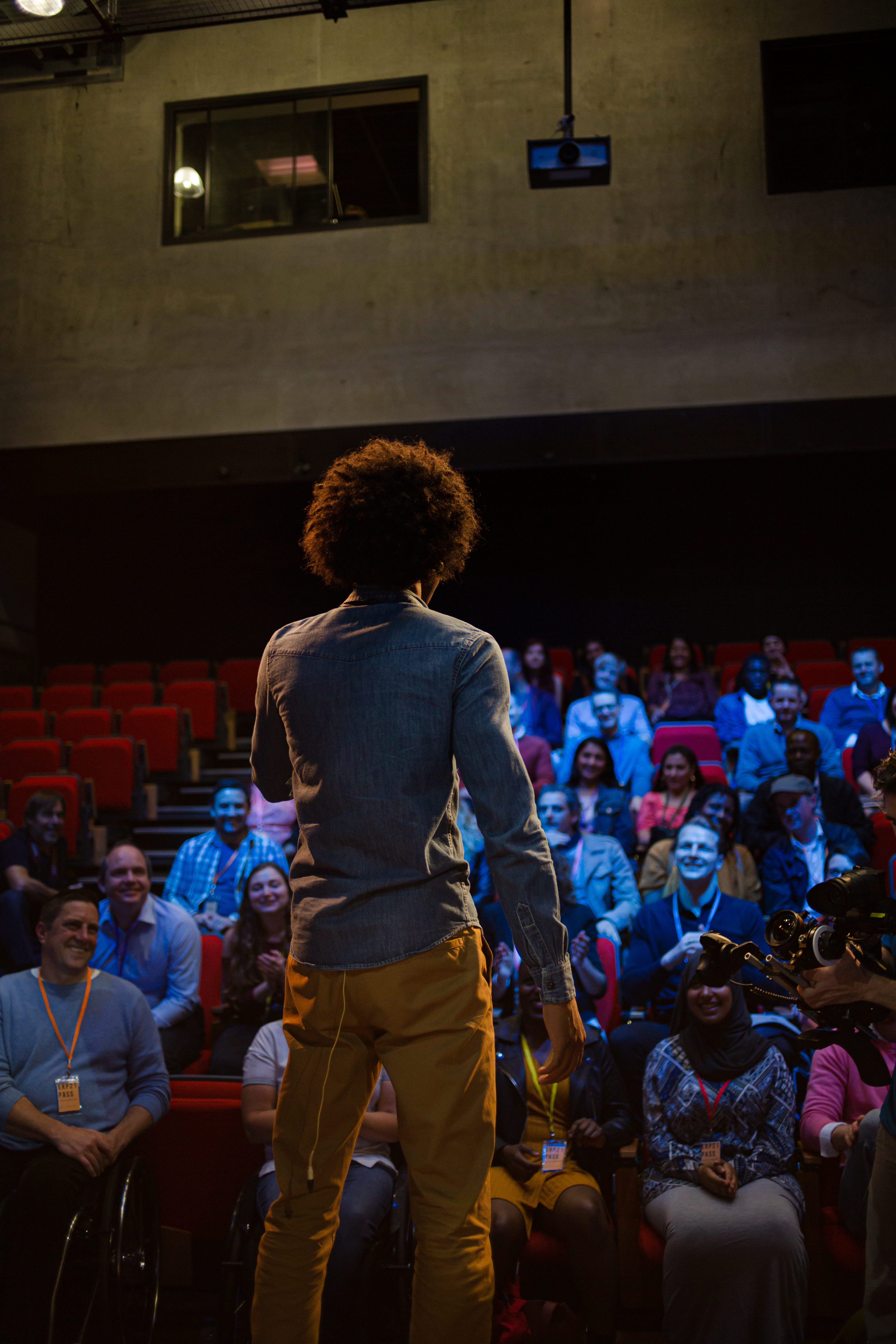 A person stands on a stage facing an audience seated in red chairs in an auditorium.