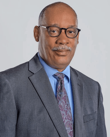 Professional headshot Attorney John Elmore, a black man with short, dark hair, dark-rimmed glasses, and a salt-and-pepper moustache wearing a grey suit, a light blue shirt, and a patterned tie against a light grey background.