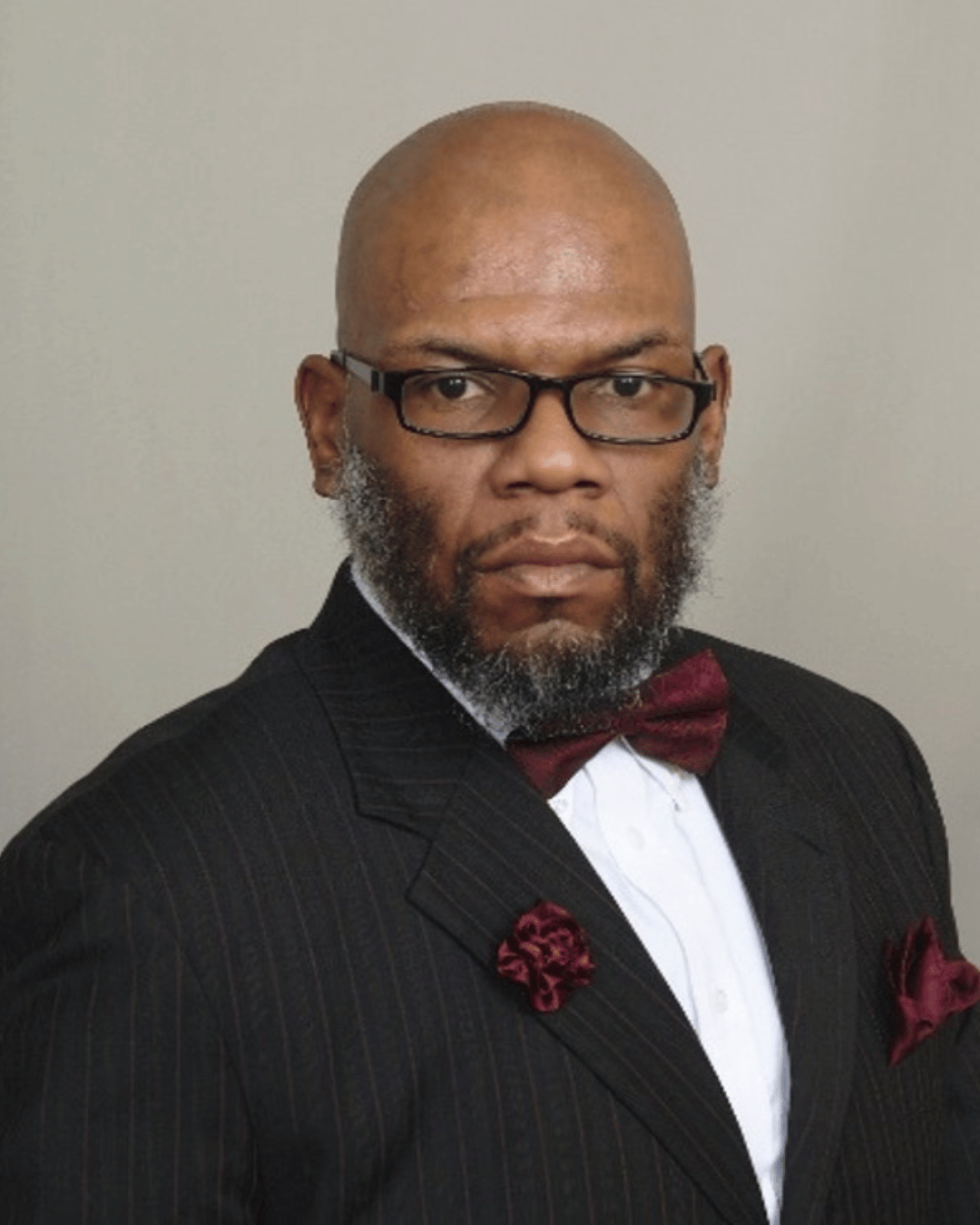 Professional headshot of Dr. Kalvin DaRonne Harvell, a bald, black man with dark-rimmed glasses and a dark beard wearing a dark, pinstriped suit, a white shirt, and a burgundy bow tie with burgundy accessories against a light gray background. 
