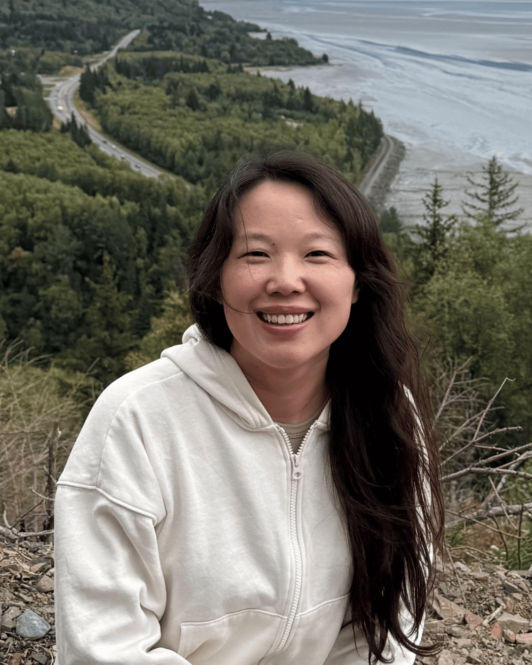 Photo of Lina Qu, a smiling Asian woman with long, dark hair wearing a cream-colored zip-up hooded sweatshirt against a backdrop of pine trees and water. 