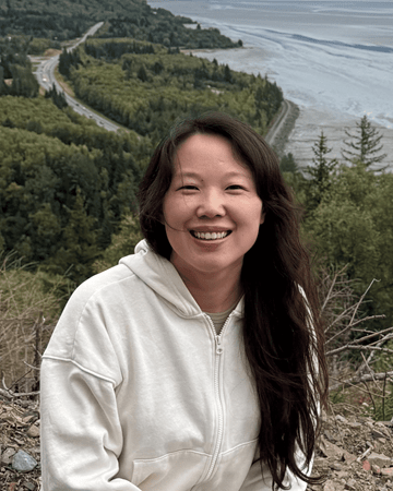 Photo of Lina Qu, a smiling Asian woman with long, dark hair wearing a cream-colored zip-up hooded sweatshirt against a backdrop of pine trees and water.