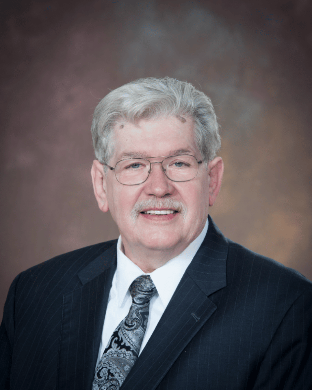Professional headshot of Richard Cunningham, a white man with white hair, a white moustache, and glasses wearing a dark, pinstriped suit, a white shirt, and a patterned tie against a brown marbled backdrop. 