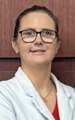 Chelsea Orito is wearing glasses with dark hair pulled up, wearing a light-colored jacket, shown from the shoulders up, placed in front a textured reddish-brown wall. 