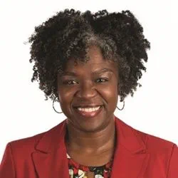 Eshe Dotson is wearing a red blazer and patterned top is shown facing the camera against a plain white background. Eshe has short, textured hair and is wearing hoop earrings.