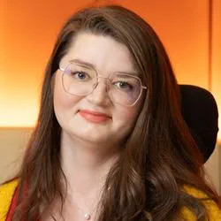 Marisa Spain is a white woman with long light brown hair and large thin rimmed geometric shaped glasses. She is wearing a red button up shirt with a mustard yellow cardigan. She is photographed smiling at the camera, sitting in her power wheelchair against a lit background with an orange-yellow gradient.