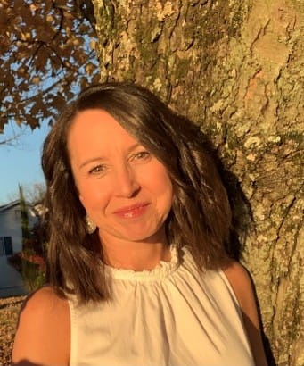 Tracie DeFreitas is standing outdoors beside a large tree trunk, illuminated by warm sunlight. Tracie has shoulder-length dark hair and is wearing a sleeveless light-colored top. Autumn leaves and a house are visible in the background.