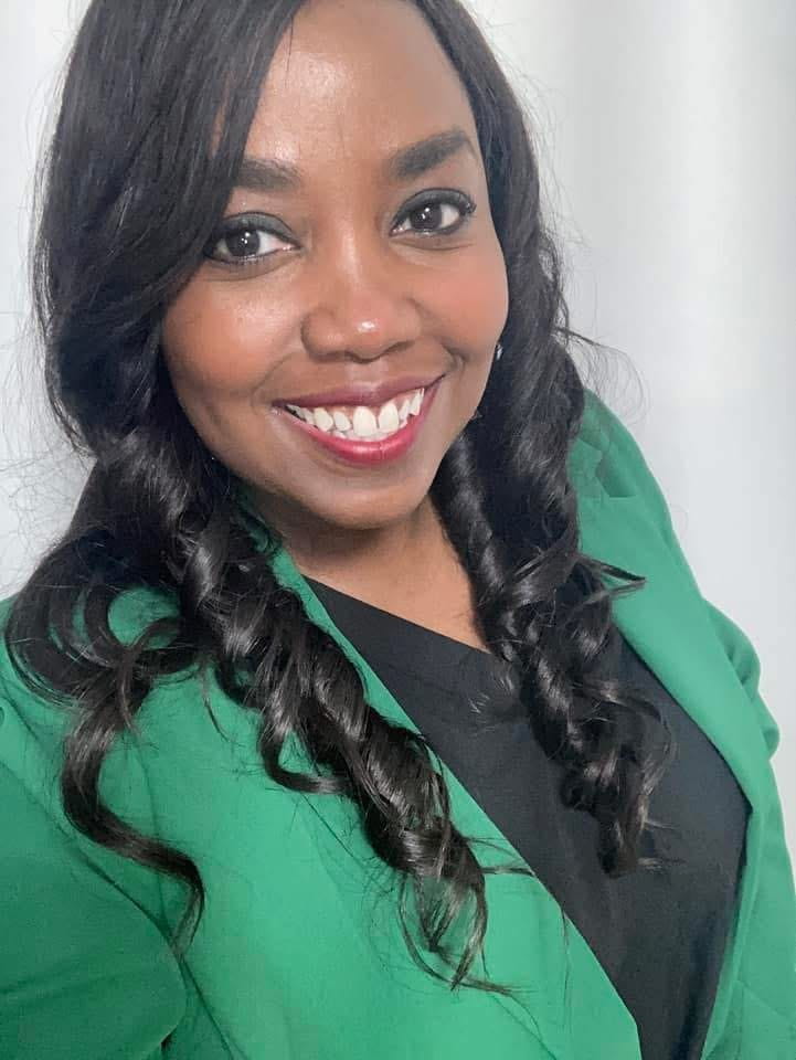 Tiara McKenzie, a dark-skinned, black female with cascading curls, is smiling while wearing red lipstick and a black blouse underneath a green blazer. She is photographed in front of a very pale, grey background. 