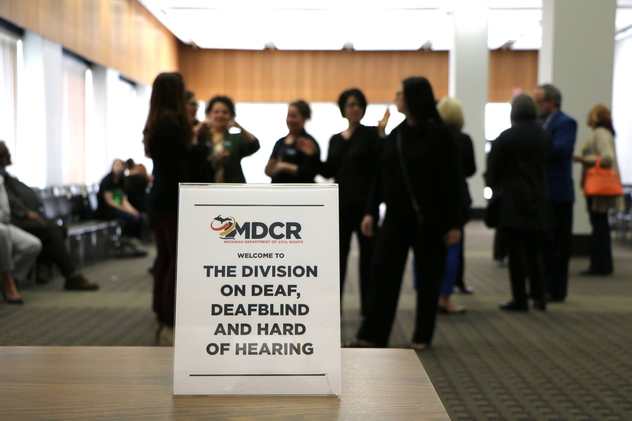 A photo of a sign with text "MDCR. Michigan Department of Civil Rights, welcome to the Division on Deaf, DeafBlind, and Hard of Hearing. In the background, people stand around talking following a meeting.