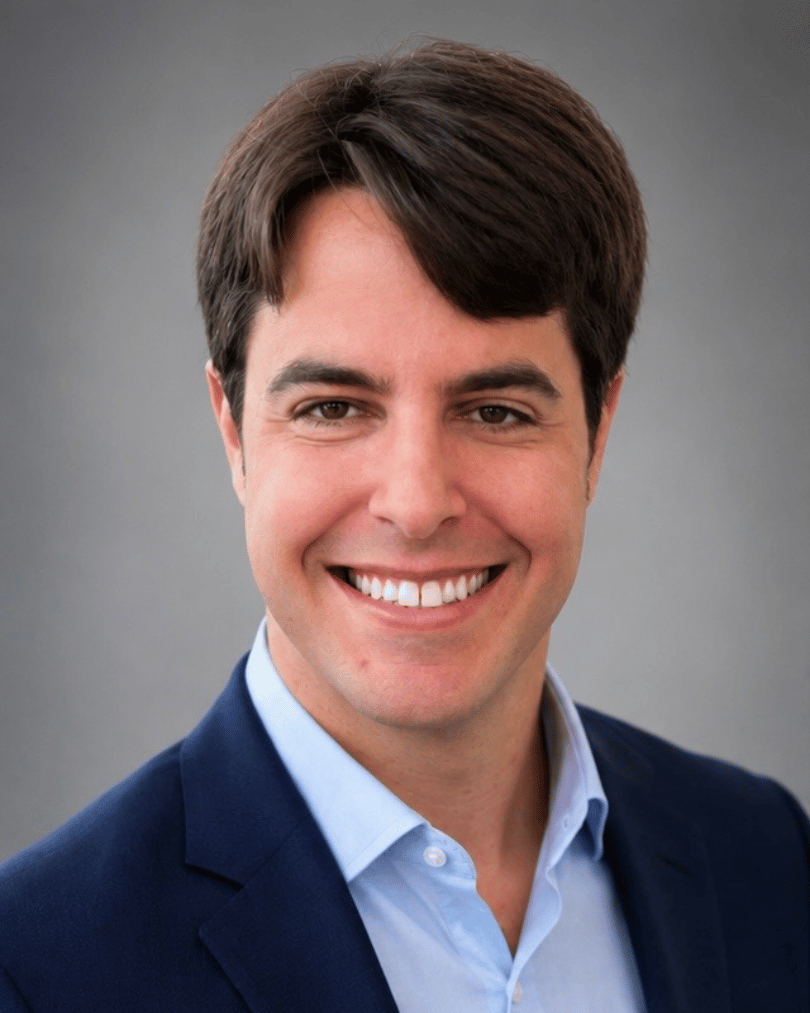 Image of Dylan Secord, a person with short, dark hair wearing a navy blue suit over a light blue shirt with a gray background