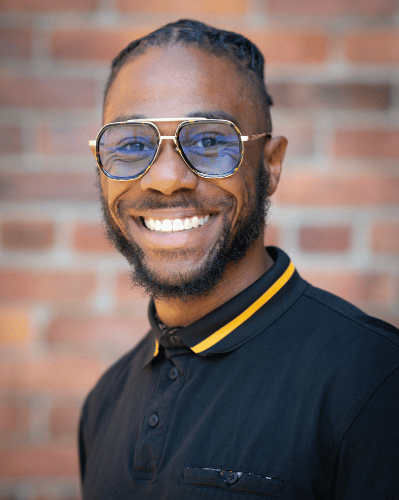 A photo of Theodore Dorsette, a person with a protective hairstyle, glasses, and a black polo shirt against a brick wall