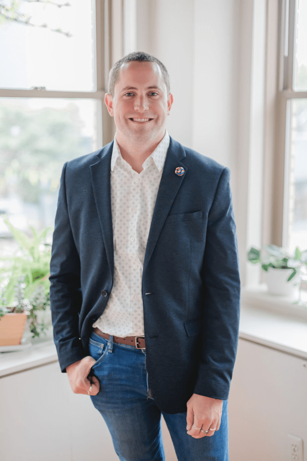 A photo of a man wearing a blue sportcoat, jeans and a white collared shirt. He is standing in a corner with two bright windows on either side. 