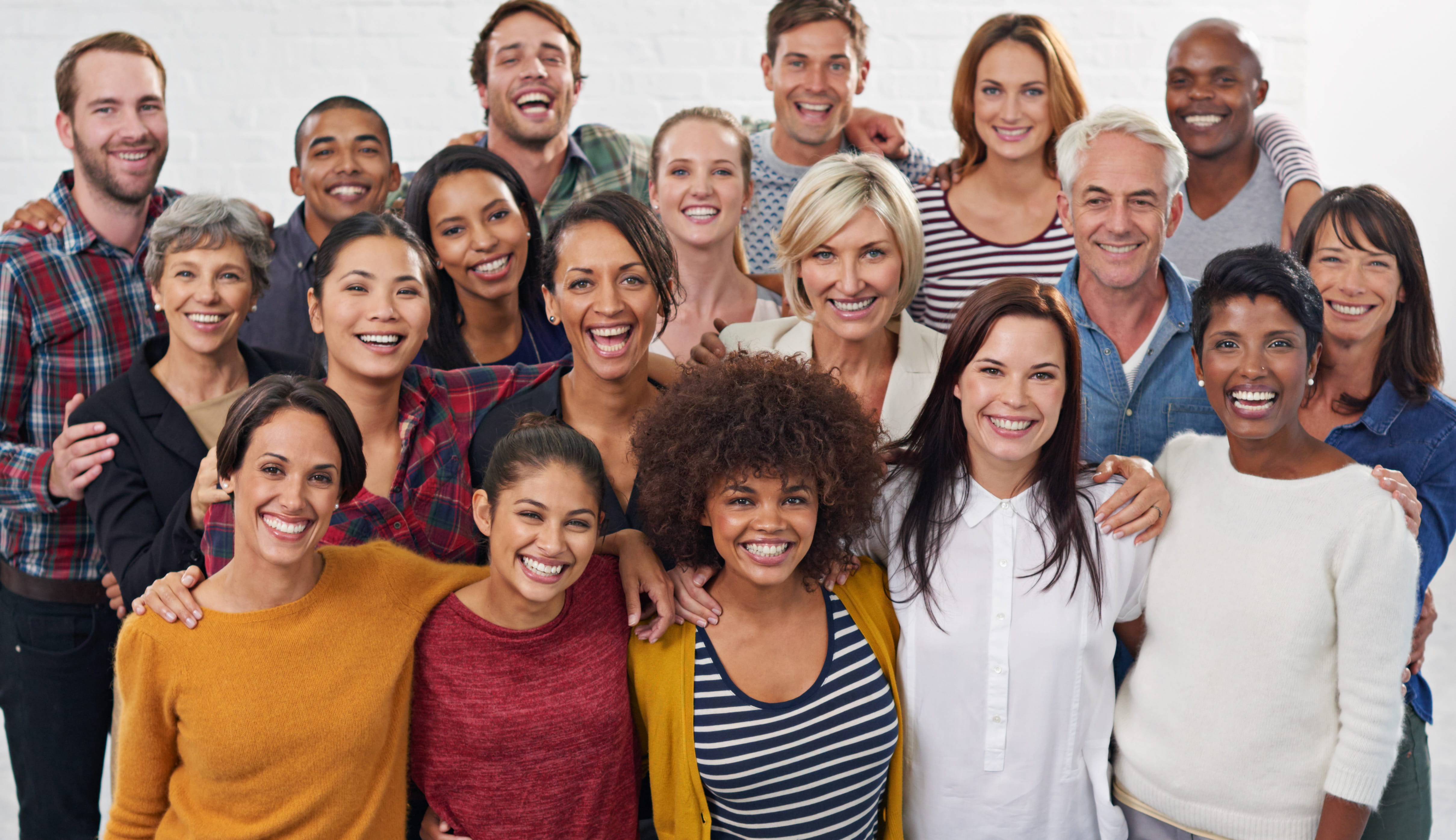 A group of people ranging in age, ethnicity and race stand with arms around shoulders smiling at the camera. The background is neutral. 