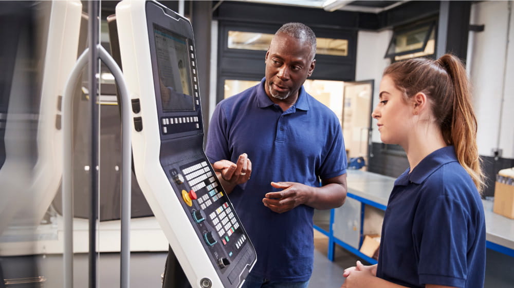 African American male instructor provides assistance to female high school student at industrial computer panel.