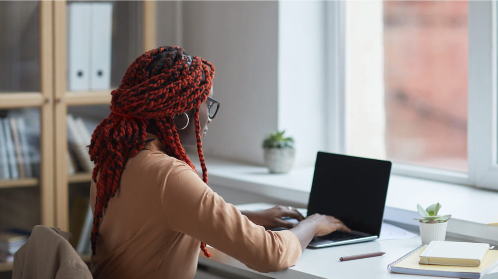 African American female high school student with red braids seated at desk working on laptop computer.